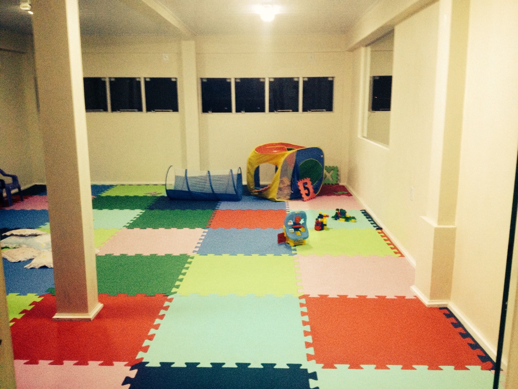 Um corredor com tapetes de espuma coloridos e entrelaçados no chão, uma tenda de brincar e uma pequena área de recreação com brinquedos.