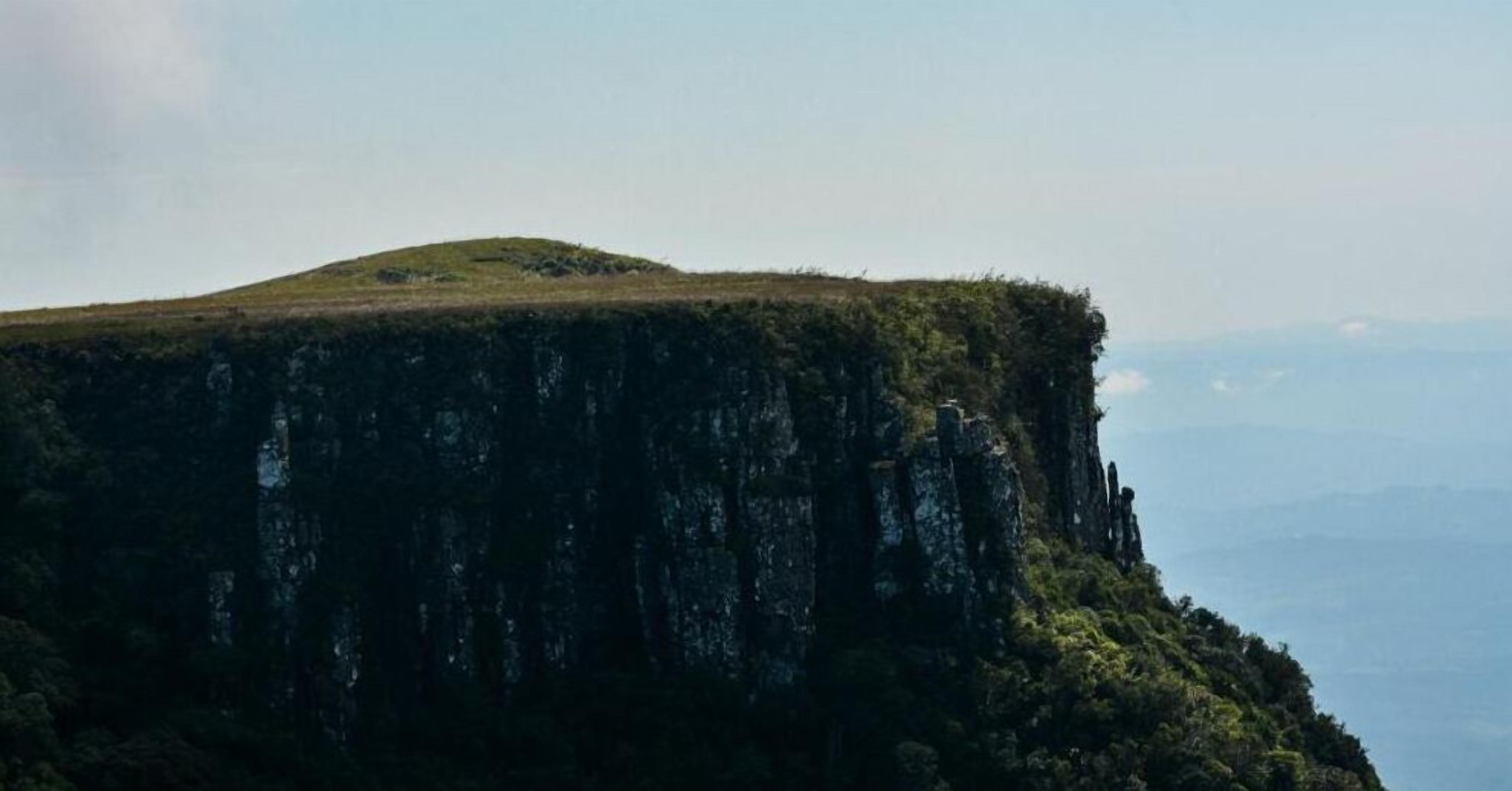 A high cliff with a grassy summit and dense forest at the base.