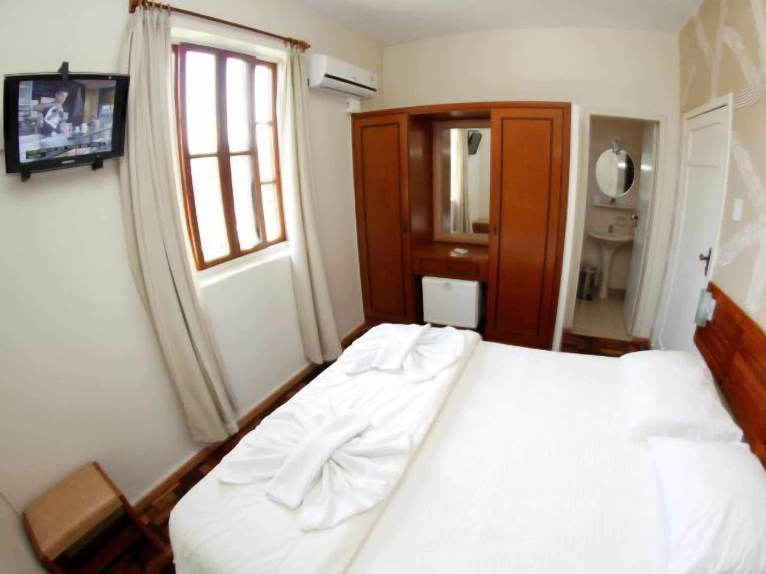 Une chambre d’hôtel avec un lit, un petit bureau, un miroir et une fenêtre avec des rideaux.