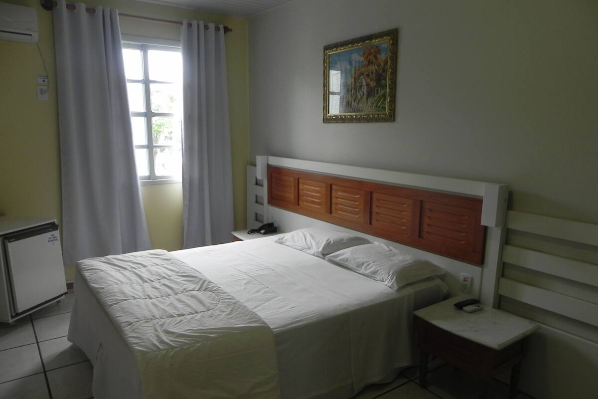 A hotel room with a bed, a bedside table, a small desk, a window with curtains, and a wall-mounted painting.