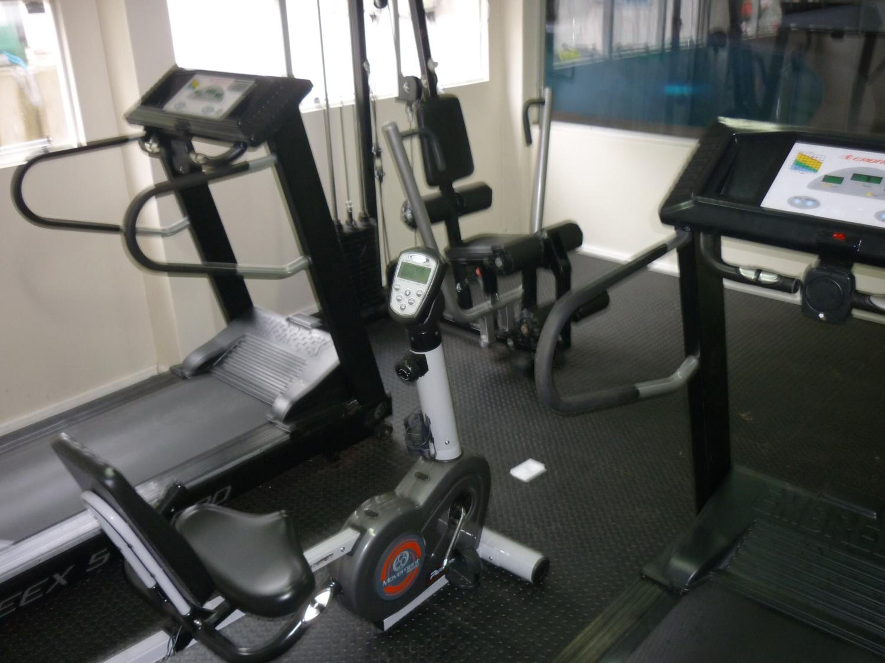 Una bicicleta estática con pantalla digital, una cinta de correr y un banco de pesas en un gimnasio.