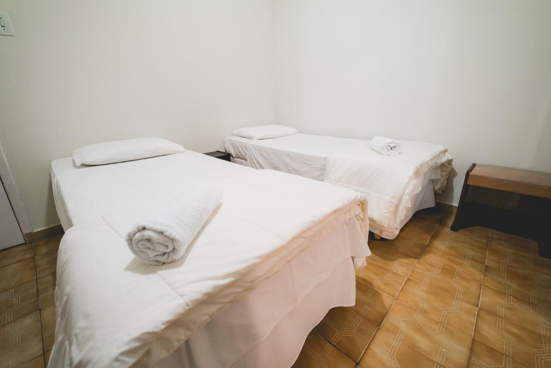 Una habitación con tres camas individuales, cada una con una colcha blanca y una toalla enrollada en blanco. El suelo es de baldosas y hay un banco de madera apoyado en la pared.
