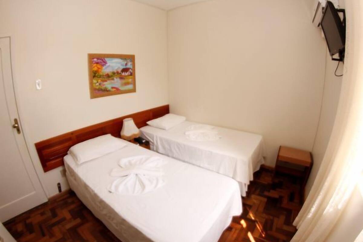 A hotel room with two single beds, a wooden headboard, a small bedside table with a lamp, and a painting on the wall.