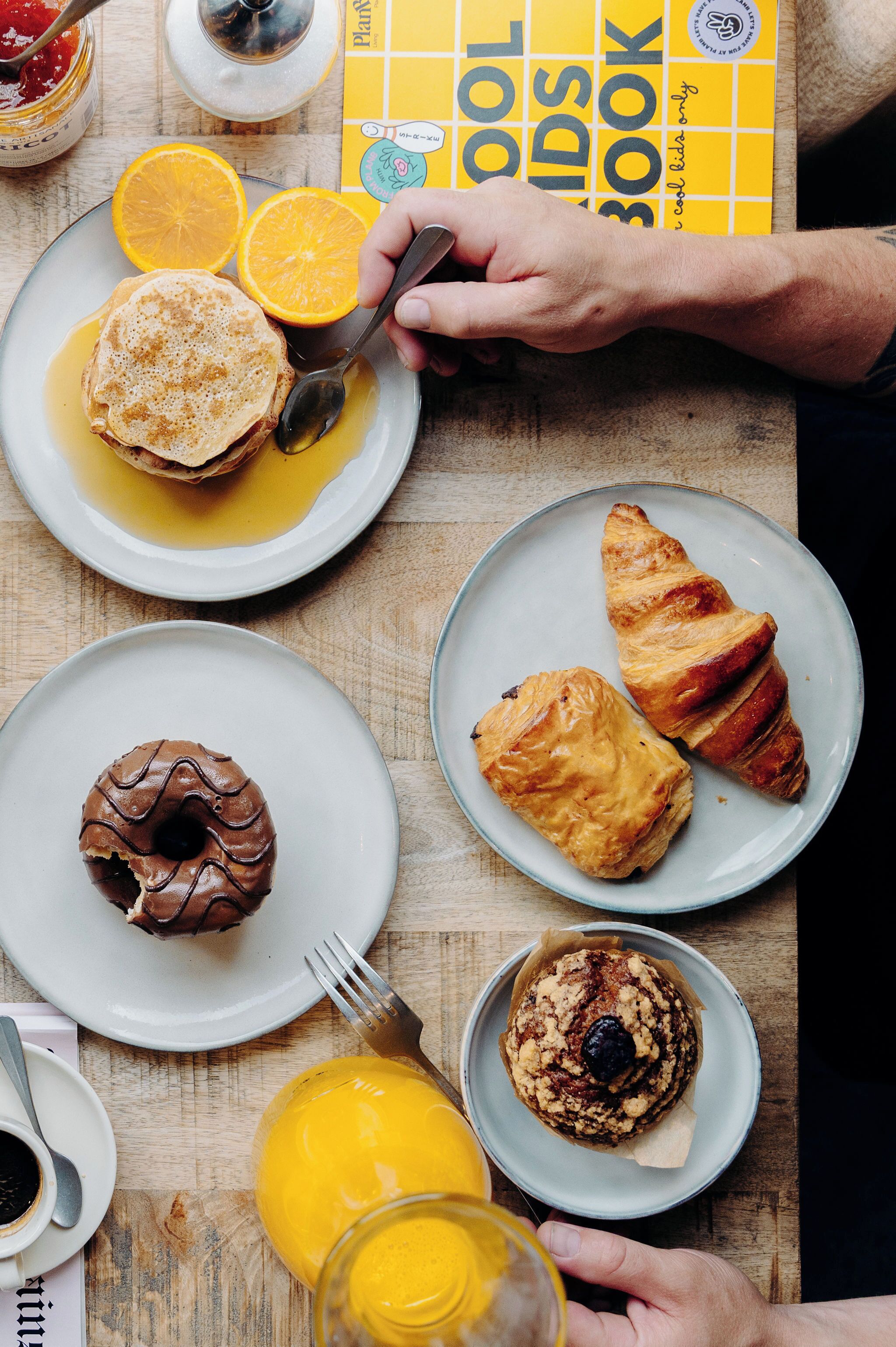 A table with breakfast items including pancakes, a donut, croissants, and a muffin, along with a glass of orange juice and a book.
