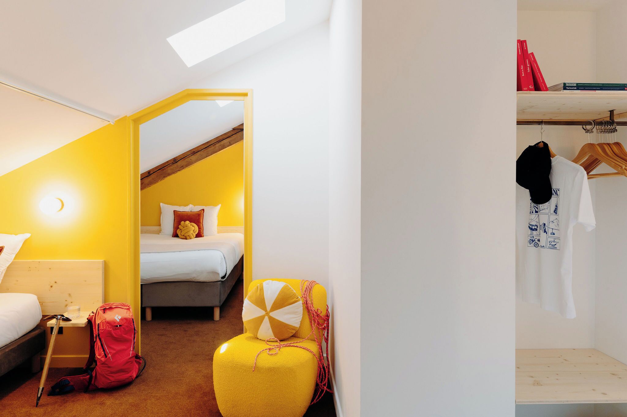 Une pièce avec un lit, une chaise jaune, un sac à dos rouge et un banc en bois.