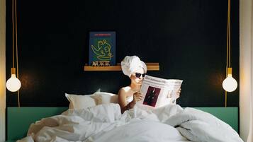 A person lying in bed reading a magazine, with a book titled 'LES DÉBUTANTS' on a shelf above.