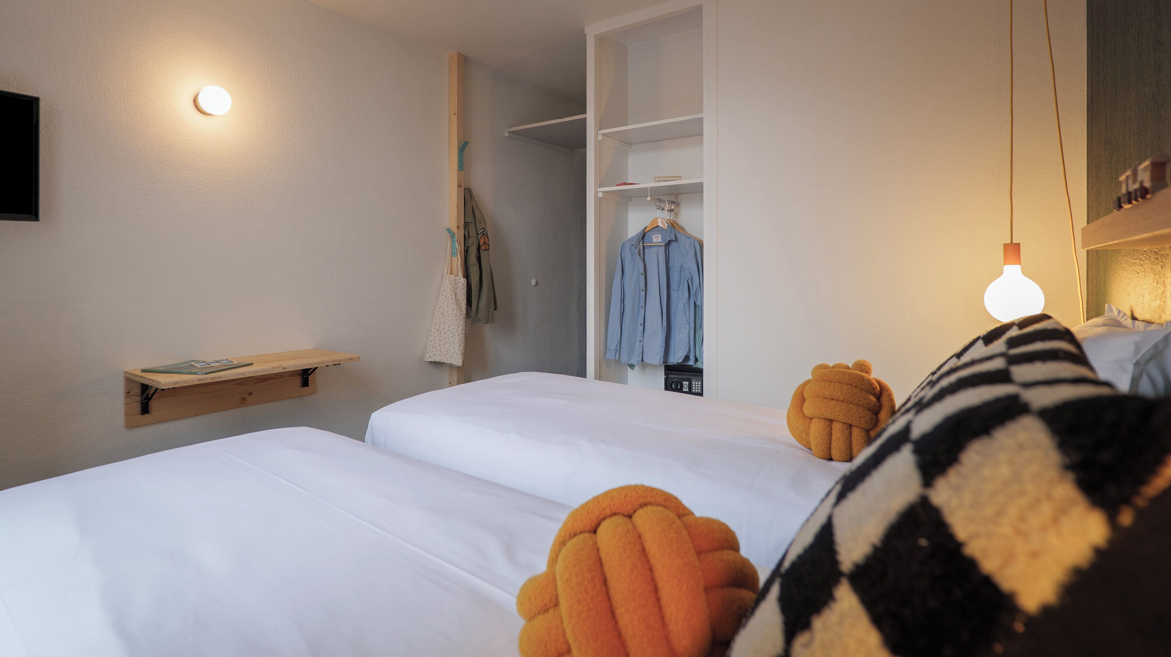 A bedroom with two beds, a wooden shelf, a hanging light, and a wardrobe with clothes.