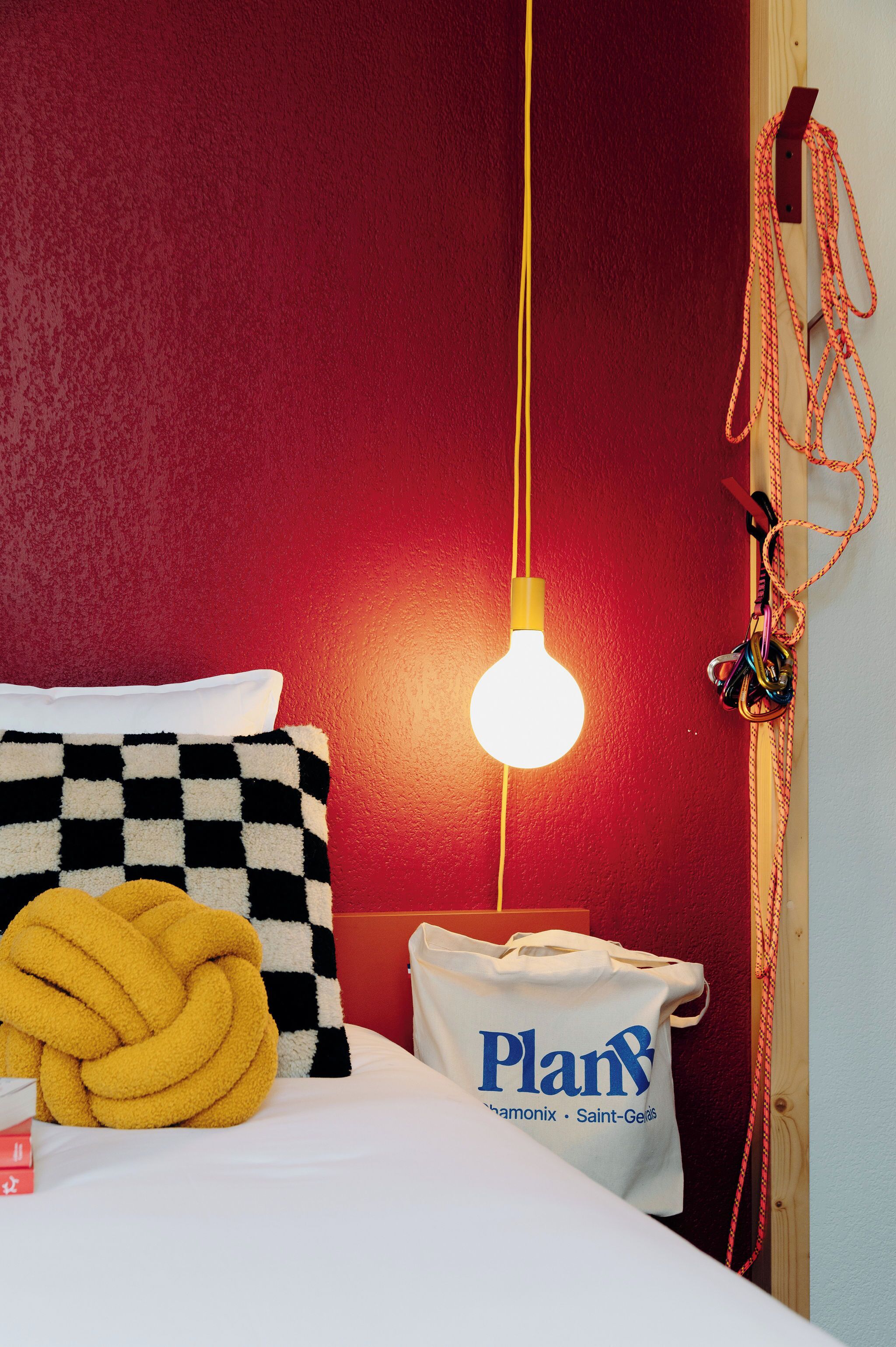 Een bed met een geruit kussen, een geel touw, een witte tas met de tekst 'Plan' en een hangende gloeilamp.