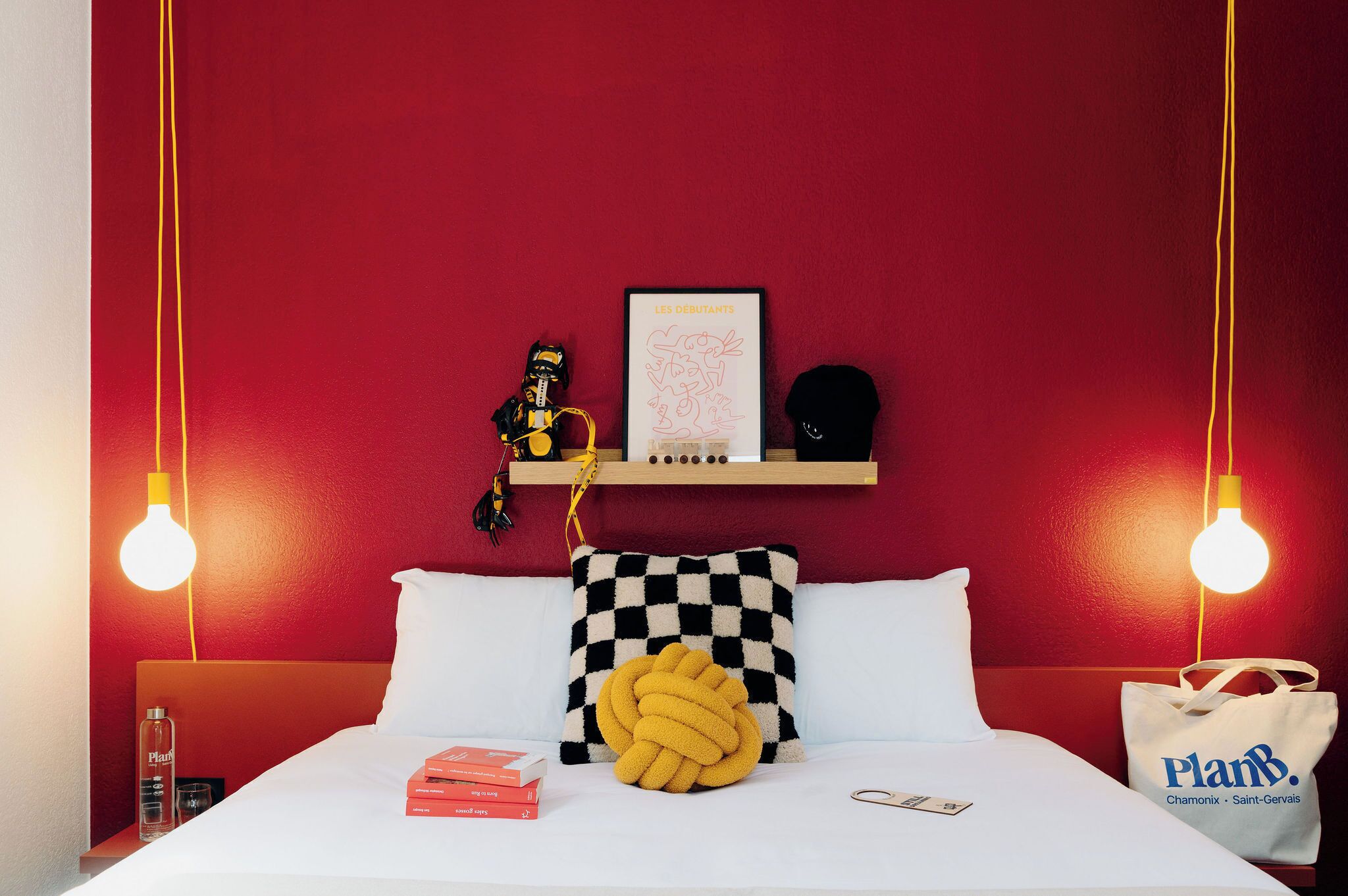 Une chambre d’hôtel avec un mur rouge, un lit avec une literie blanche, un oreiller à carreaux, un article tricoté jaune, un tableau encadré et une bouteille d’eau de la marque PlanB.