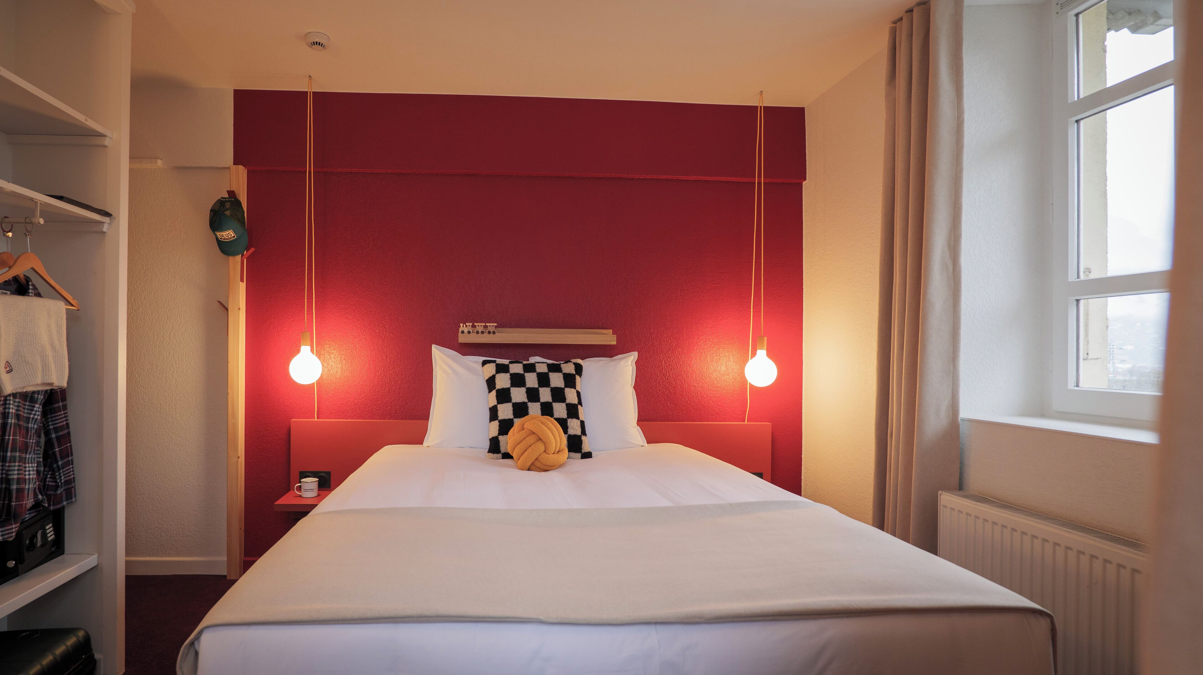 Quarto de hotel com parede de destaque vermelha, cama com roupa de cama branca, duas luminárias de cabeceira e uma janela com cortinas.