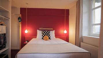 Quarto de hotel com parede de destaque vermelha, cama com roupa de cama branca, duas luminárias de cabeceira e uma janela com cortinas.