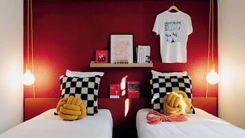 Habitación de hotel con dos camas, almohadones cuadriculados, una pared roja, un estante con libros y una camiseta, y una lámpara colgante.