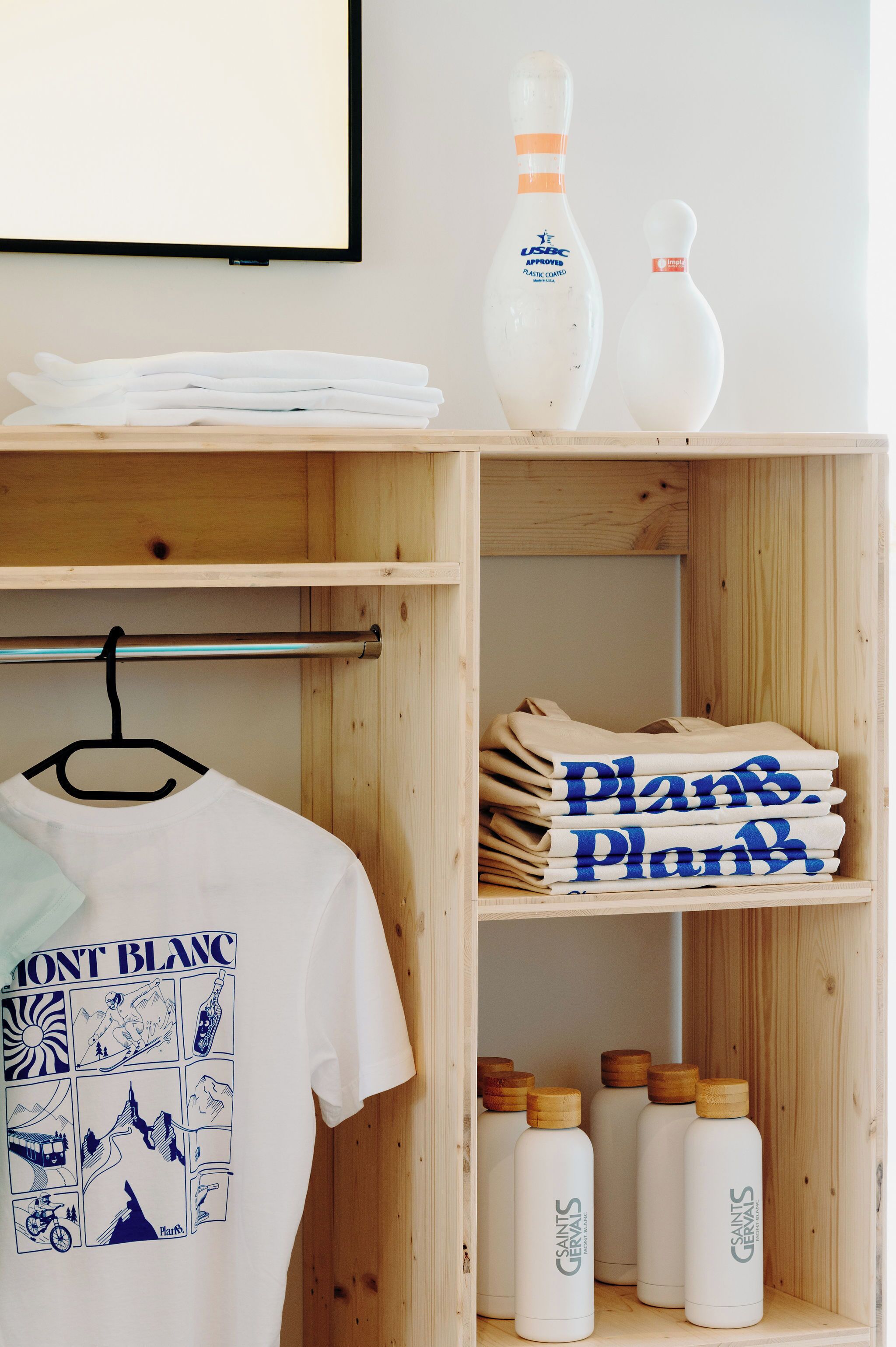 Une étagère en bois avec des serviettes pliées, un t-shirt blanc et des bouteilles d’eau de Saint-Gervais.