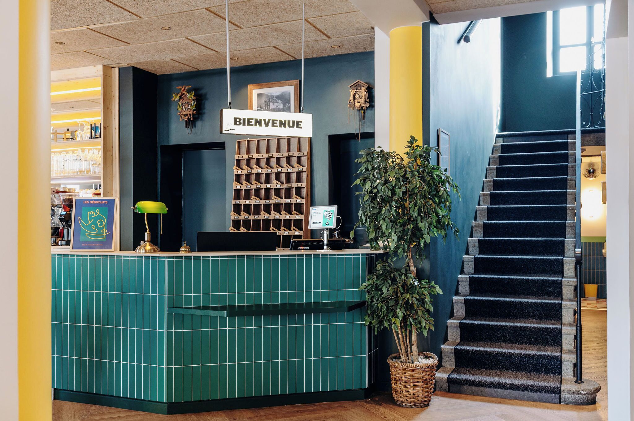 A reception area with a green tiled counter, a 'BIENVENUE' sign, a wine rack, and a staircase.