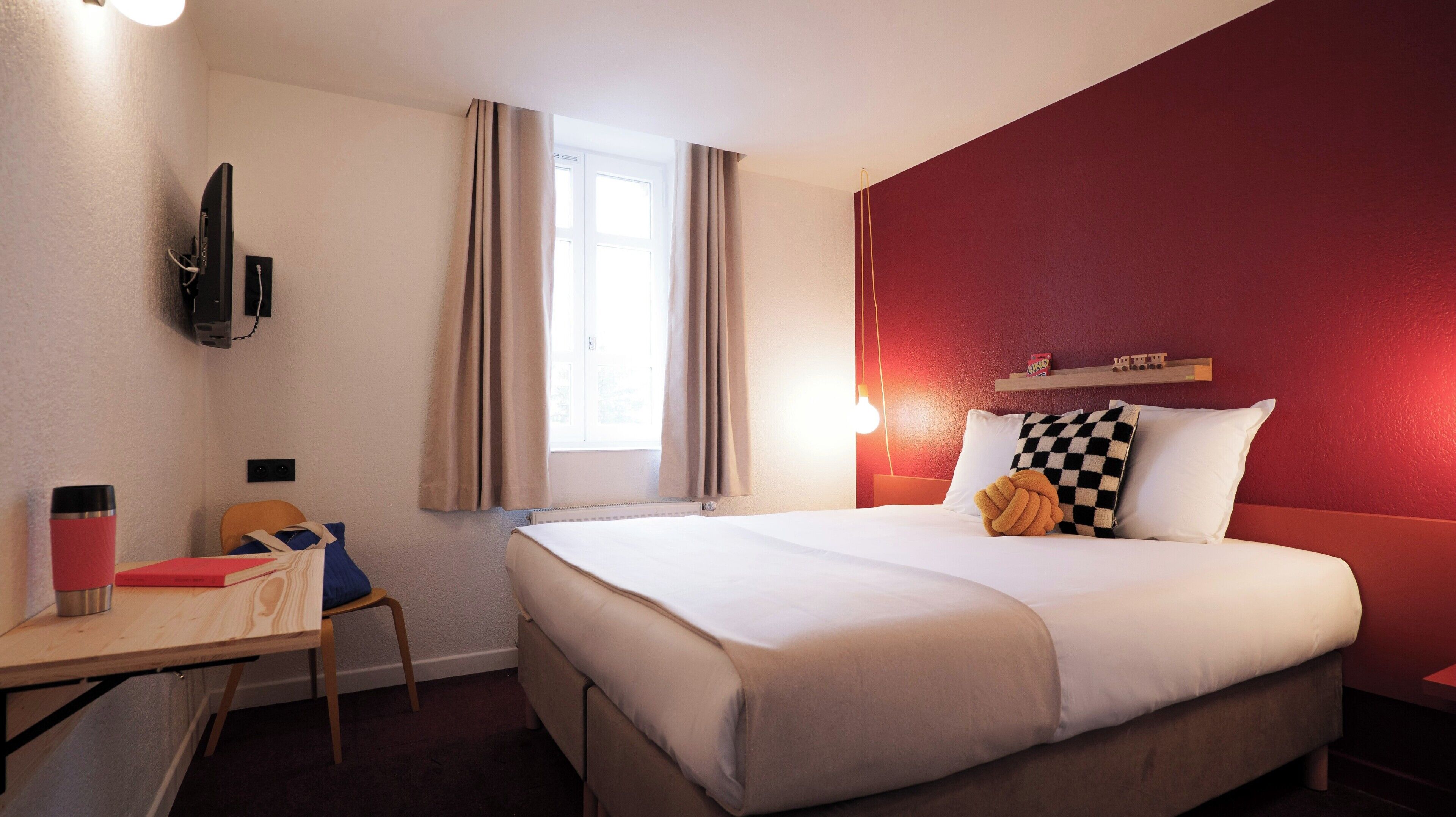 A hotel room with a bed, a desk, a chair, a TV, and a window with curtains.