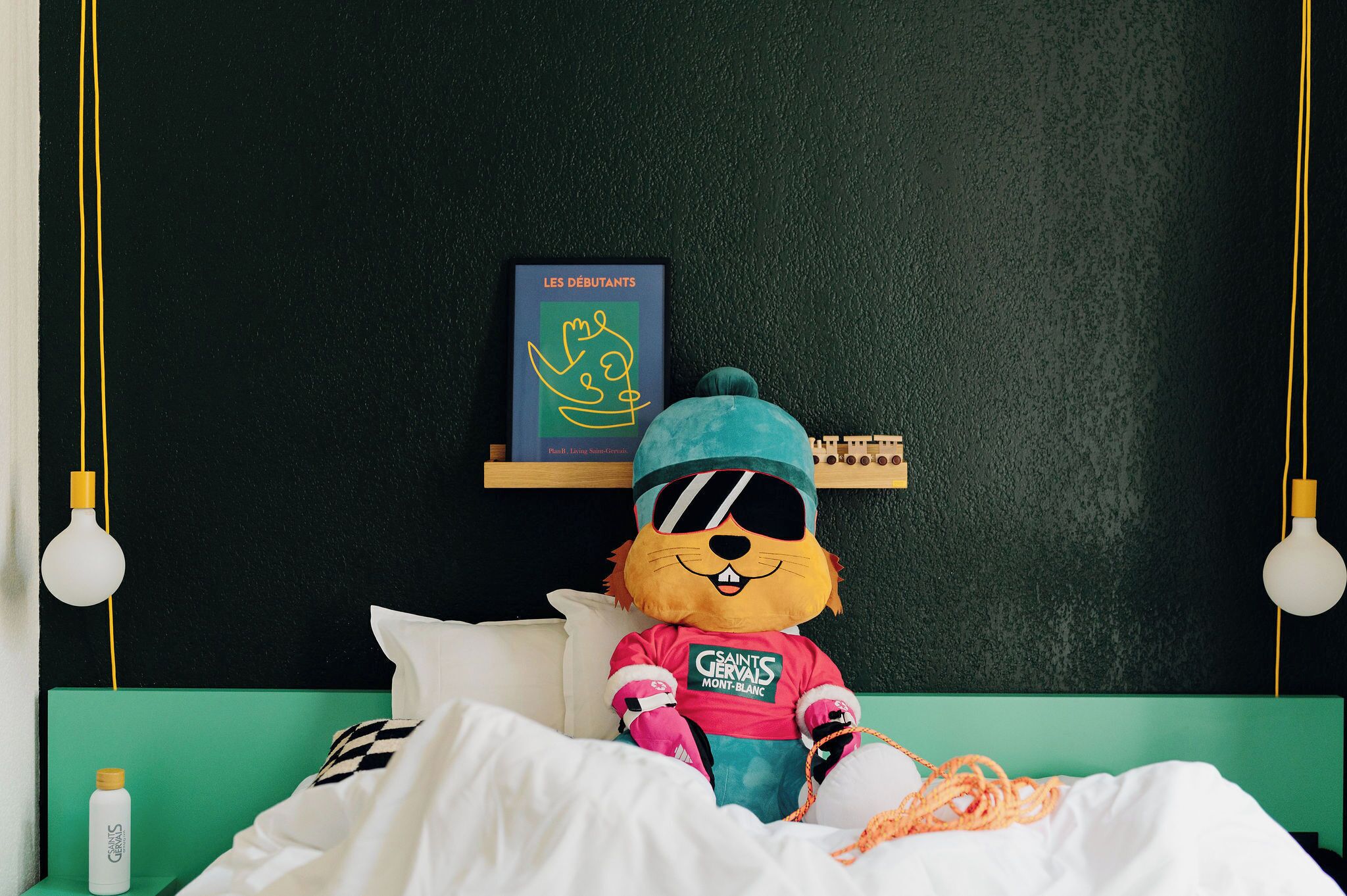Un peluche sobre una cama con cabecera verde, un libro enmarcado en una estantería y una botella en la mesita de noche.