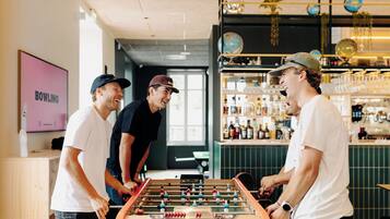 Uma área de bar com mesa de pebolim, prateleiras com garrafas e uma tela de TV exibindo "BOWLING".