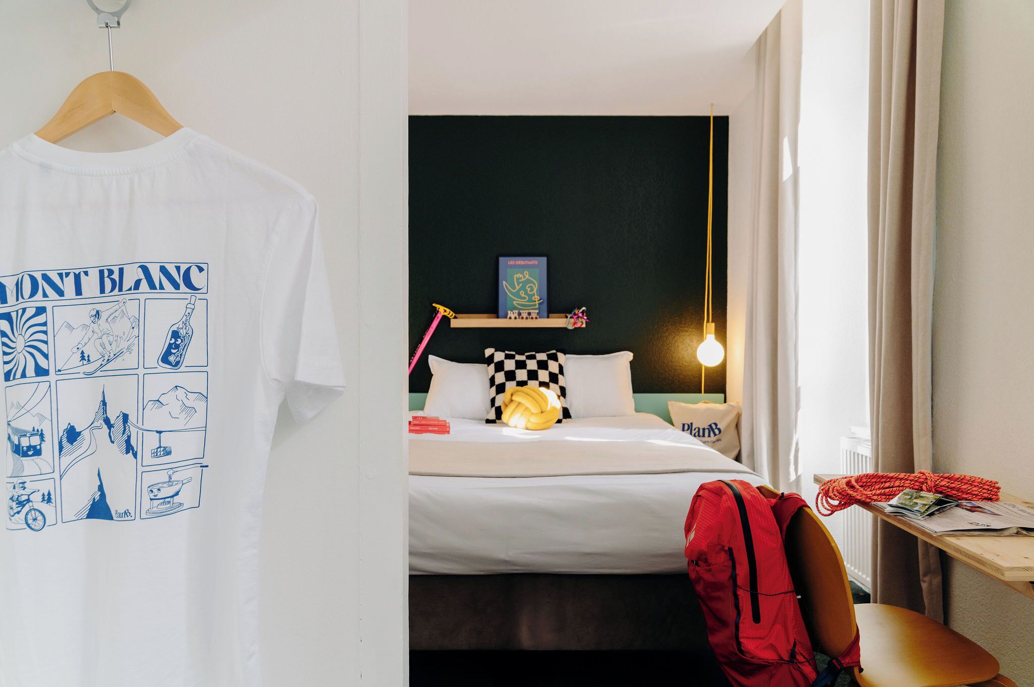 Una habitación de hotel con una cama, una mesita de noche con una lámpara, una mochila roja y una camiseta blanca colgada en una puerta.