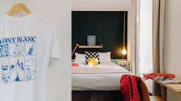 Una habitación de hotel con una cama, una mesita de noche con una lámpara, una mochila roja y una camiseta blanca colgada en una puerta.
