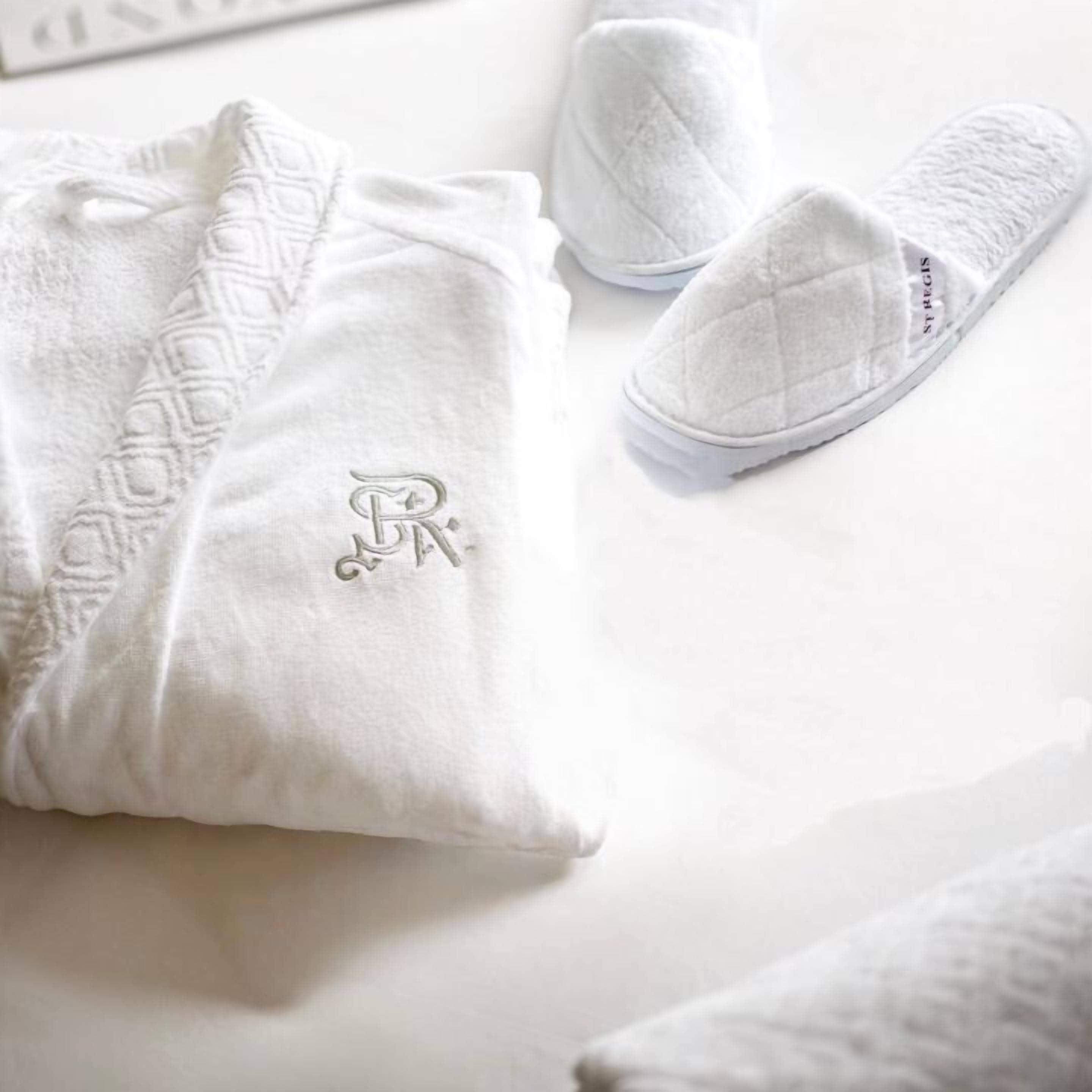 A white pillow with a monogram, a pair of white slippers, and a white blanket on a white surface.