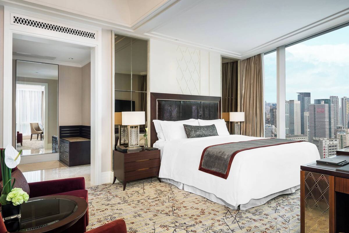 A hotel room with a large bed, a desk, a chair, a nightstand, and a view of the city.