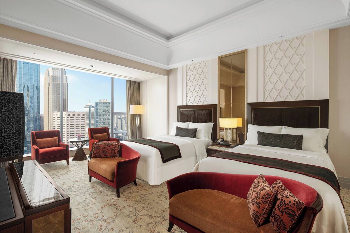 A hotel room with two beds, a sitting area with chairs, a TV, and a large window with a city view.