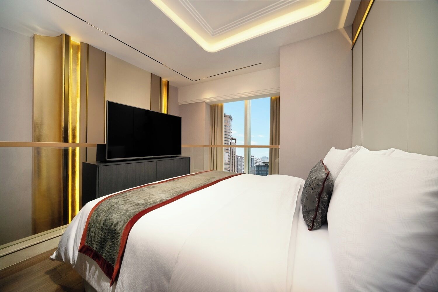 Una habitación de hotel moderna con una cama grande, un televisor de pantalla plana montado sobre un mueble oscuro y vistas a la ciudad a través de amplias ventanas.