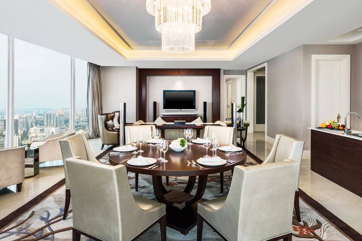 A modern hotel room with a dining area, a large chandelier, a flat-screen TV, and a city view.