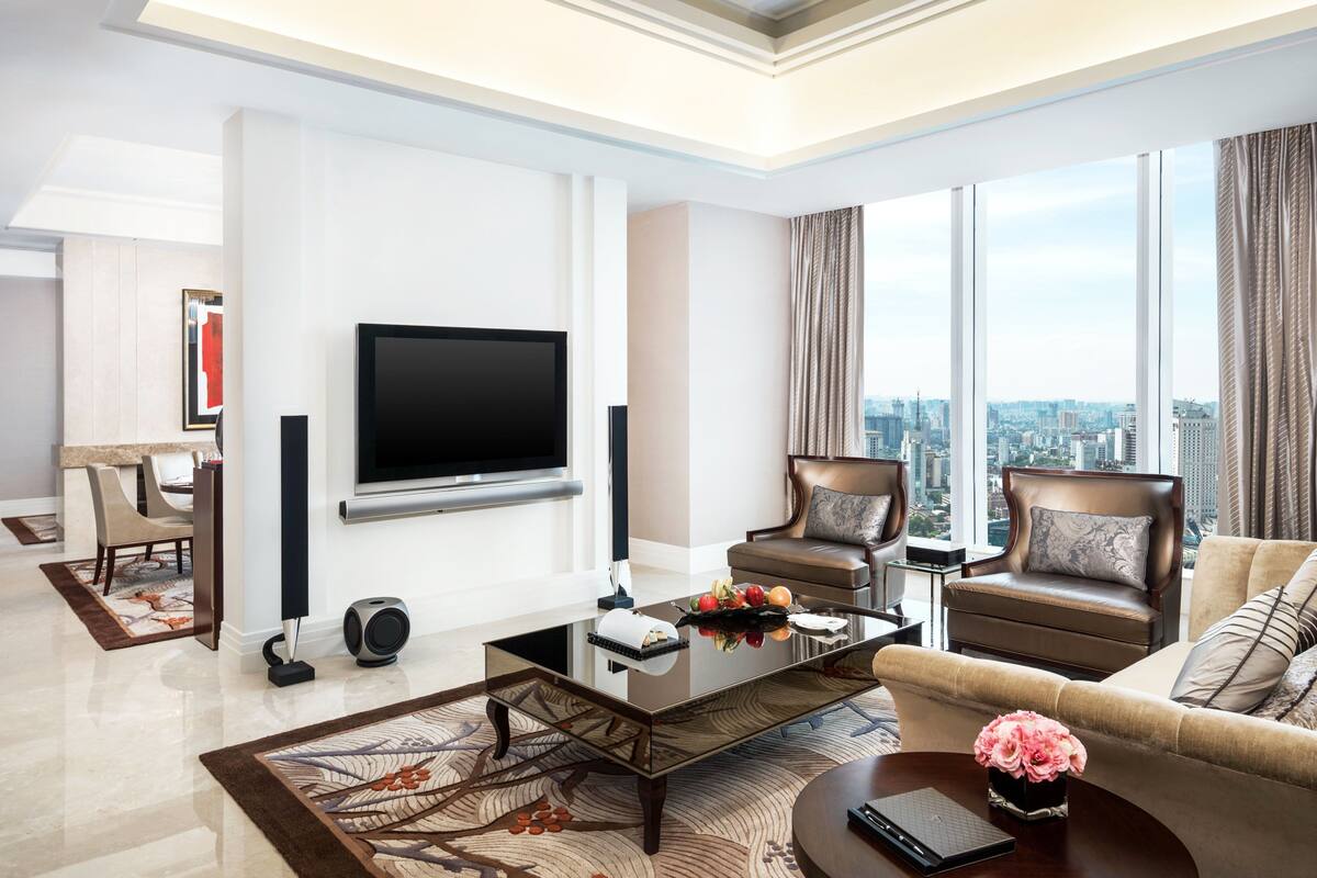 A modern living room with a flat-screen TV, two leather armchairs, a coffee table, and a city view through large windows.