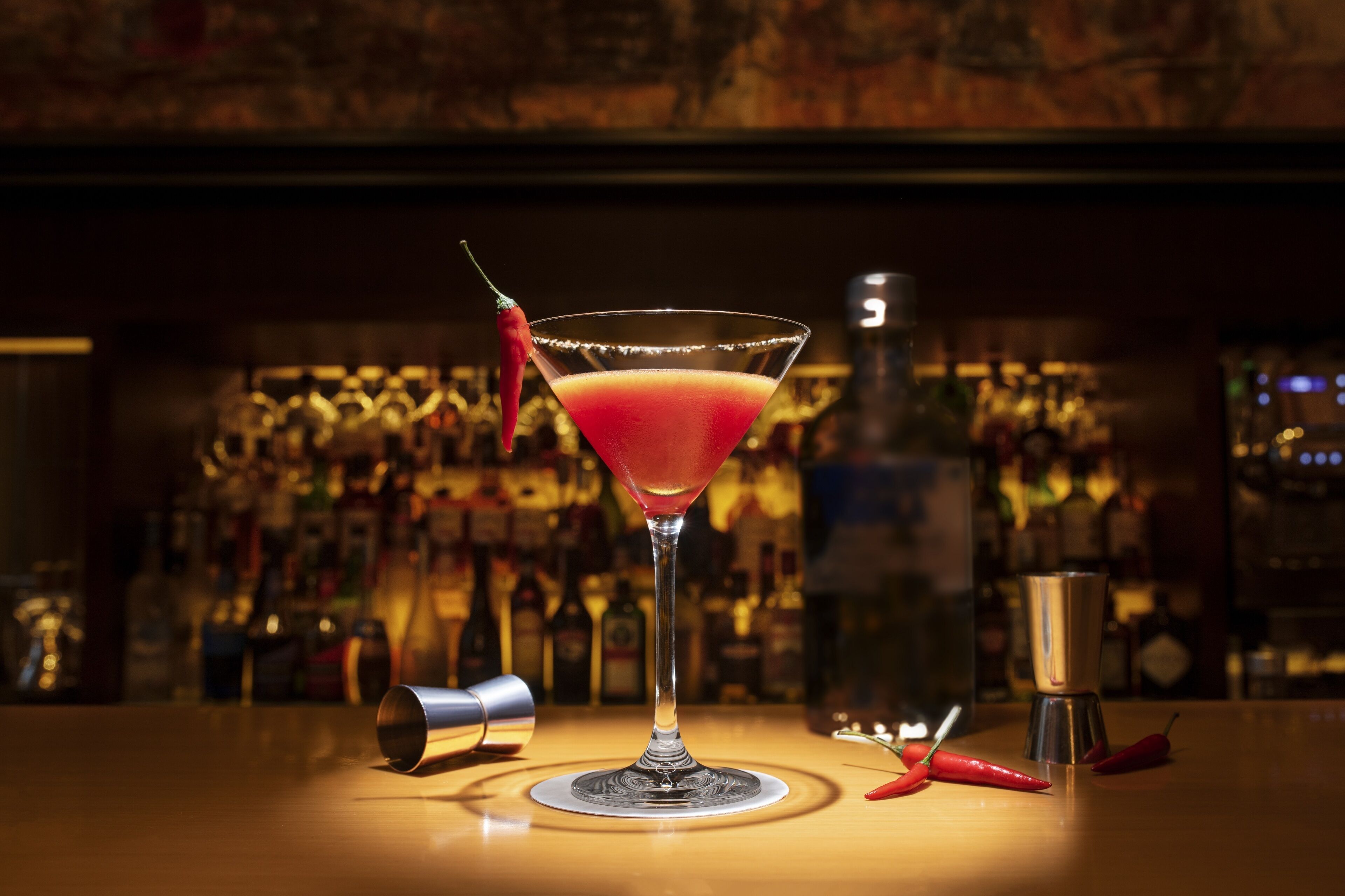 Un verre à martini contenant une boisson rouge, un shaker à cocktails et deux piments rouges sur le comptoir d’un bar.