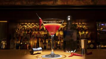 Un verre à martini contenant une boisson rouge, un shaker à cocktails et deux piments rouges sur le comptoir d’un bar.