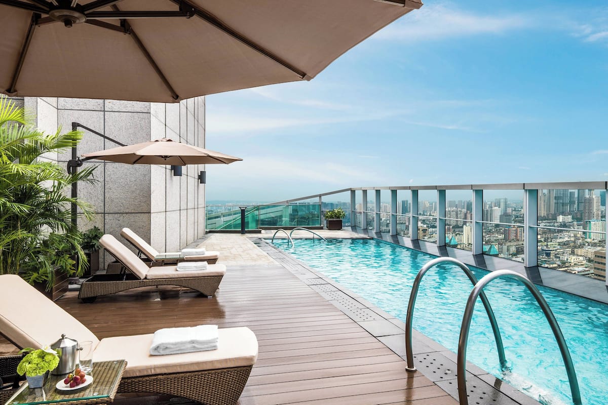 A rooftop infinity pool with lounge chairs and umbrellas, overlooking a cityscape.
