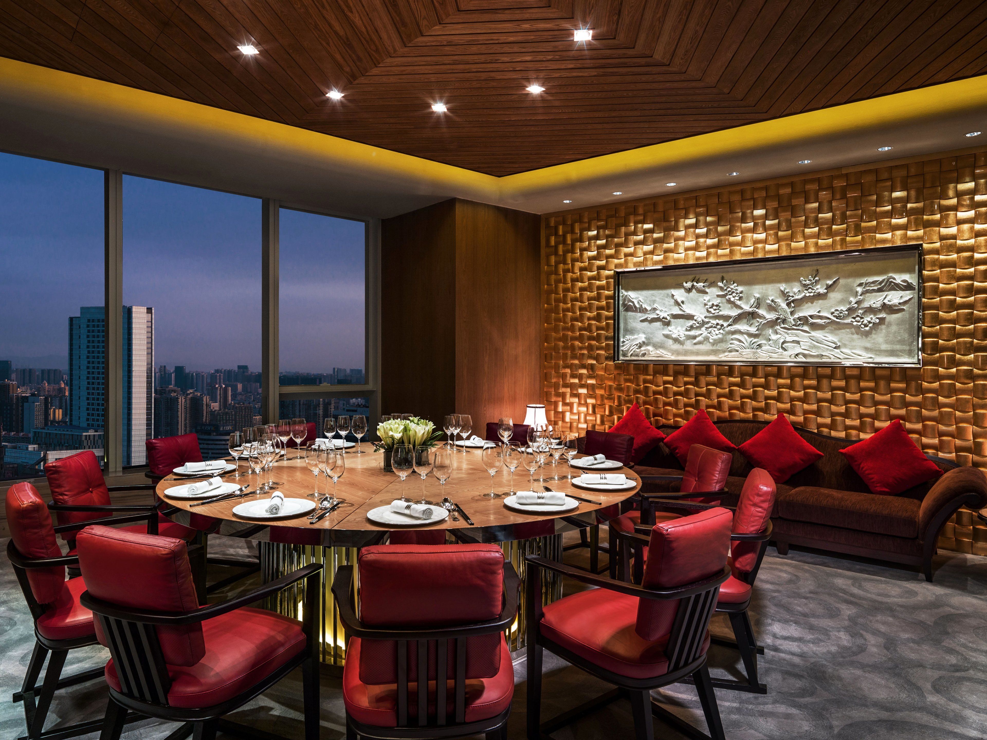 Un coin repas avec une table ronde dressée pour le repas, entourée de chaises rouges, et une vue sur la ville à travers de grandes fenêtres.