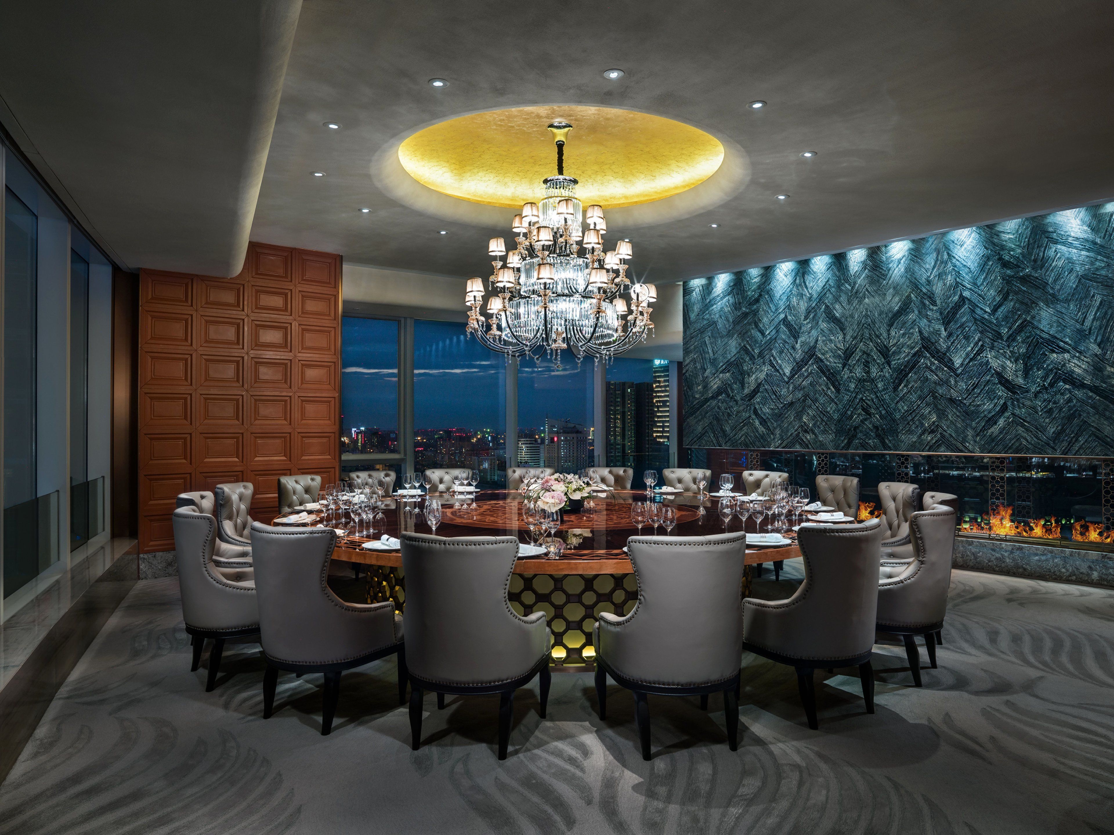 Un comedor lujoso con una gran mesa redonda dispuesta para ocho comensales, rodeada de sillas de cuero. Un candelabro imponente cuelga del techo, y la habitación cuenta con un amplio ventanal con vistas a la ciudad.