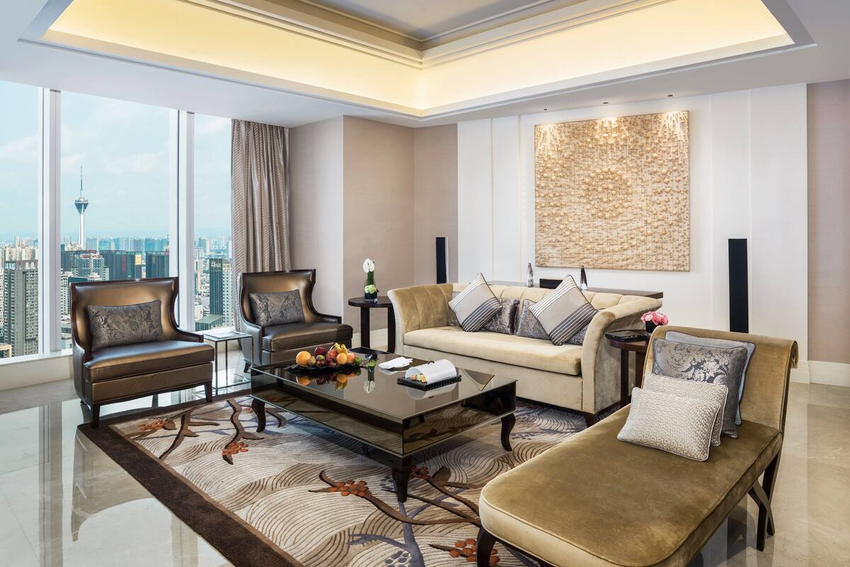A spacious living room with a modern design, featuring a sofa, armchairs, a coffee table, and a large window offering a city view.
