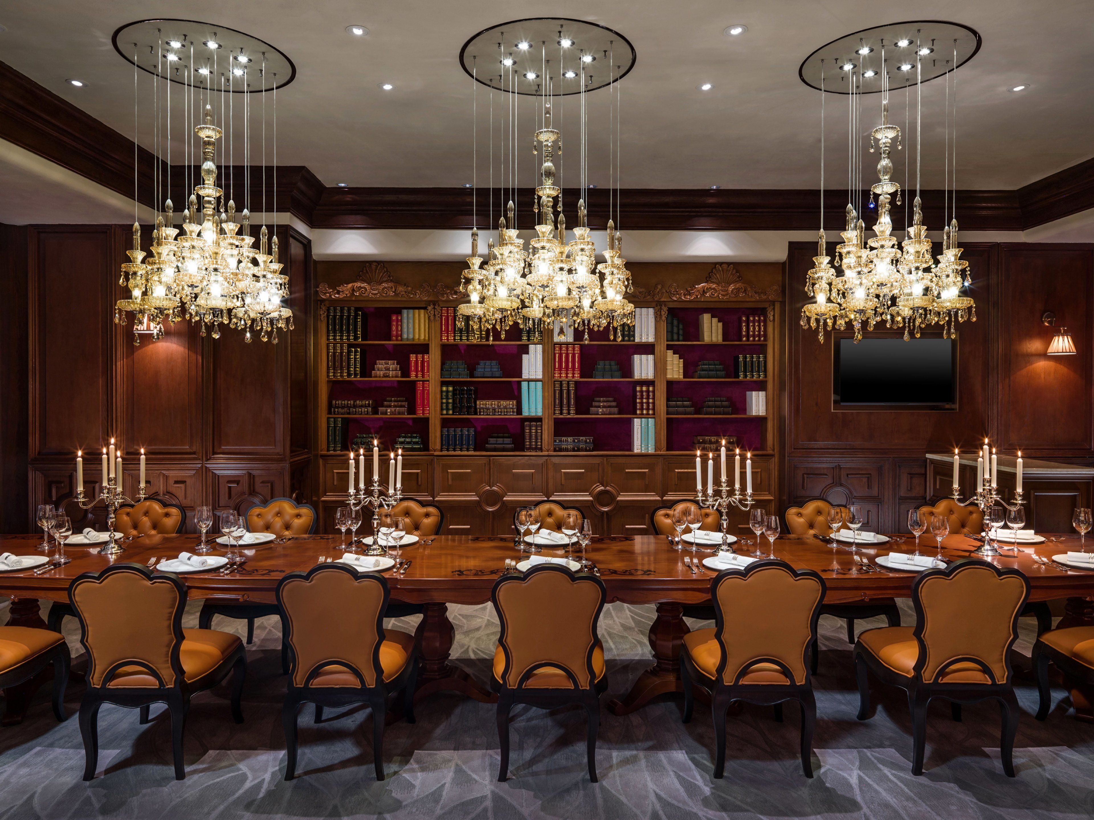 A formal dining room with a long table set for eight, surrounded by leather chairs, with three chandeliers hanging from the ceiling and a bookshelf filled with books in the background.