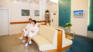 A couple sitting on a beige sofa in a well-lit room with a wooden floor, a glass table with a vase of flowers, and framed artwork on the walls. - Hotel Reza (Frantiskovy Lazne)