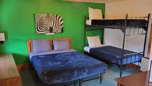A room with two beds, one with a wooden headboard and the other with a metal frame. There is a green wall with a zebra painting, a ceiling light, and a small table with a chair.