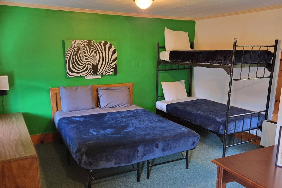 A room with two beds, one with a wooden headboard and the other with a metal frame. There is a green wall with a zebra painting, a ceiling light, and a small table with a chair.