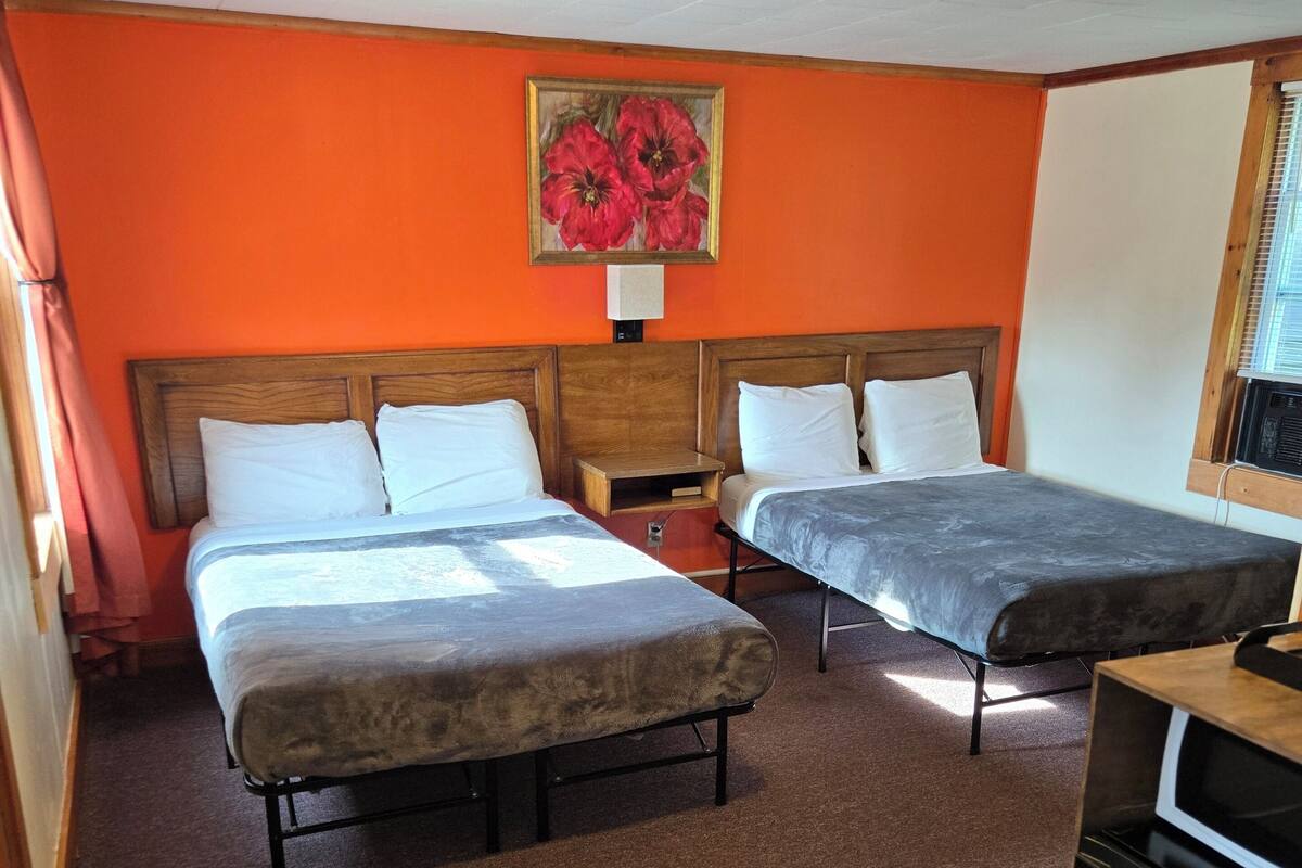 A hotel room with two beds, a wooden headboard, a painting of flowers, a small wooden nightstand, and a kitchenette area.