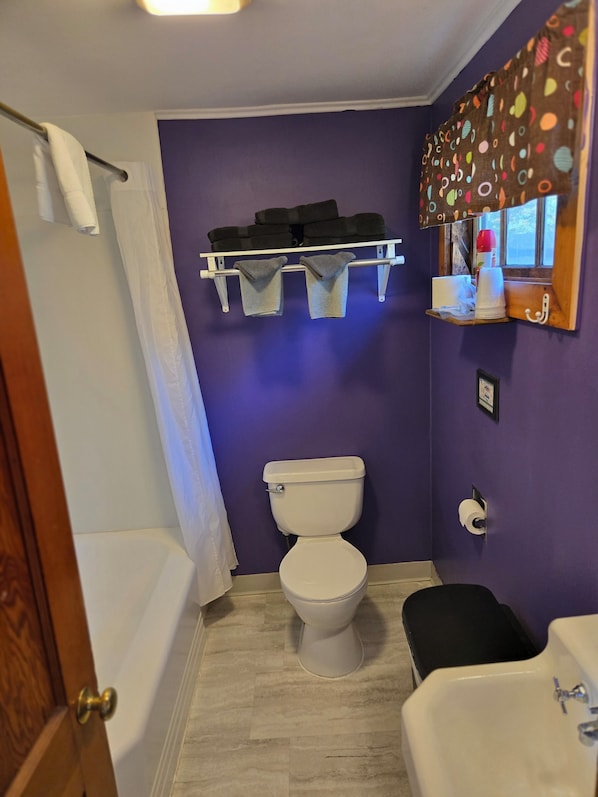 A bathroom with a white toilet, a bathtub, a sink, and a shelf with towels and toiletries.