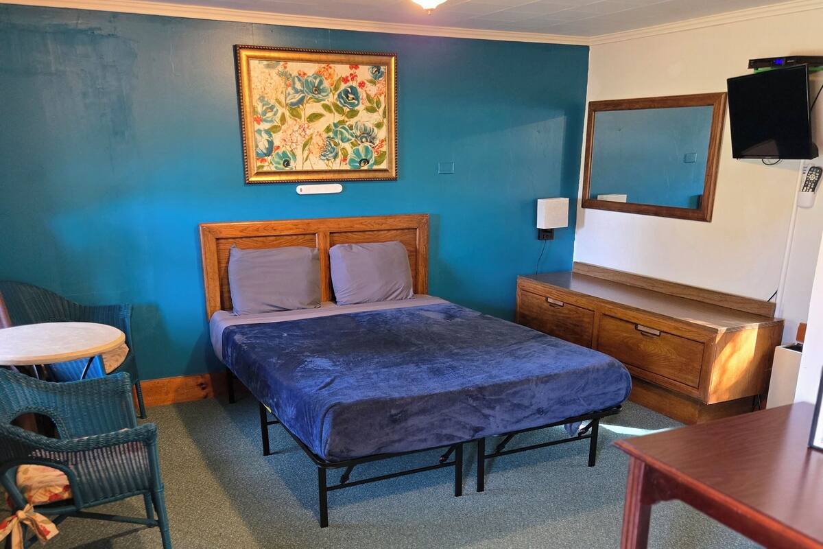 A bedroom with a bed, a dresser, a chair, a small table, and a painting on the wall.