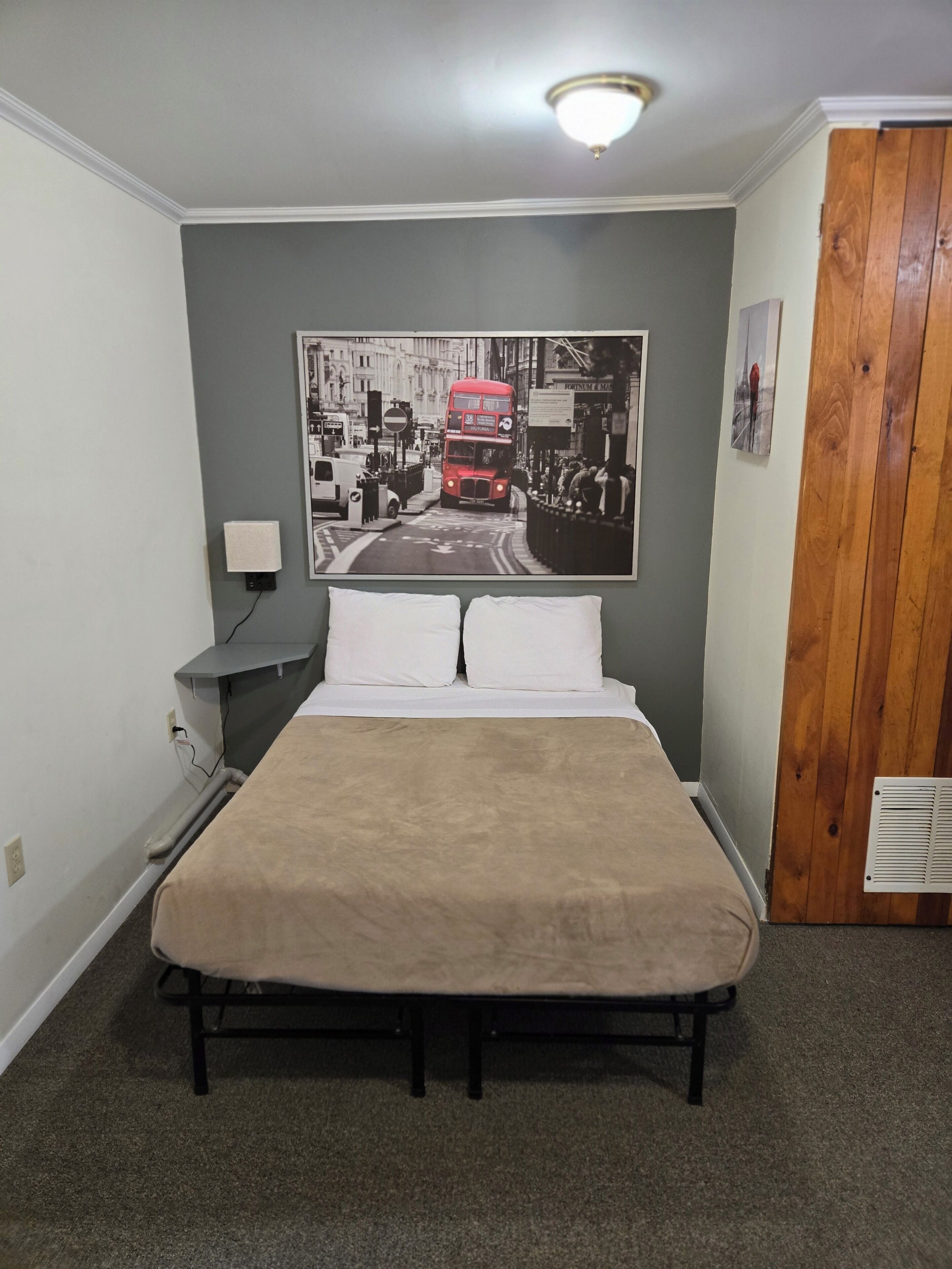 A bedroom with a bed, a nightstand, a wall-mounted lamp, a poster of a red double-decker bus, and a wooden door.