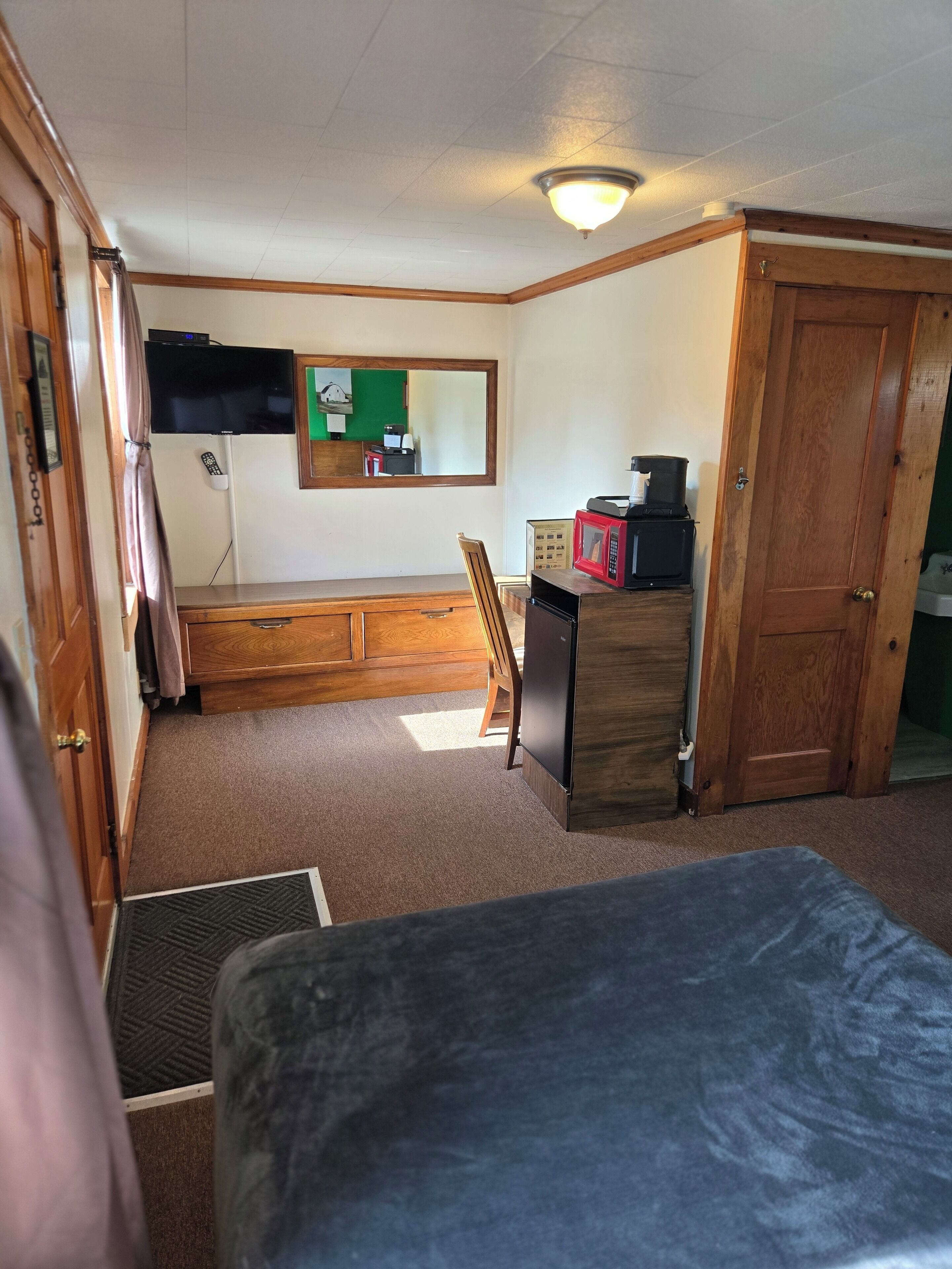 Una habitación compacta con una cama, un aparador de madera, un televisor fijado a la pared y una puerta que da a otra habitación.