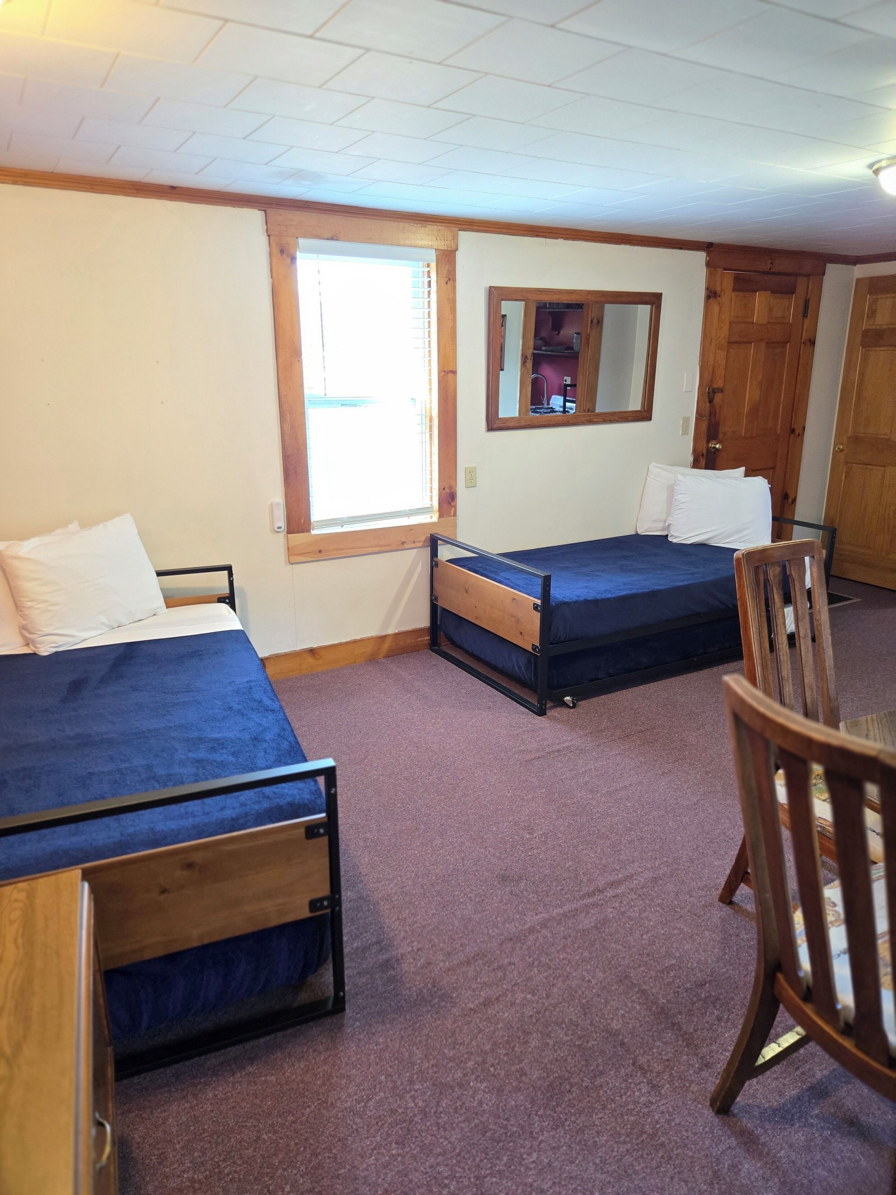 A room with two single beds, a wooden chair, a window with blinds, and a door.