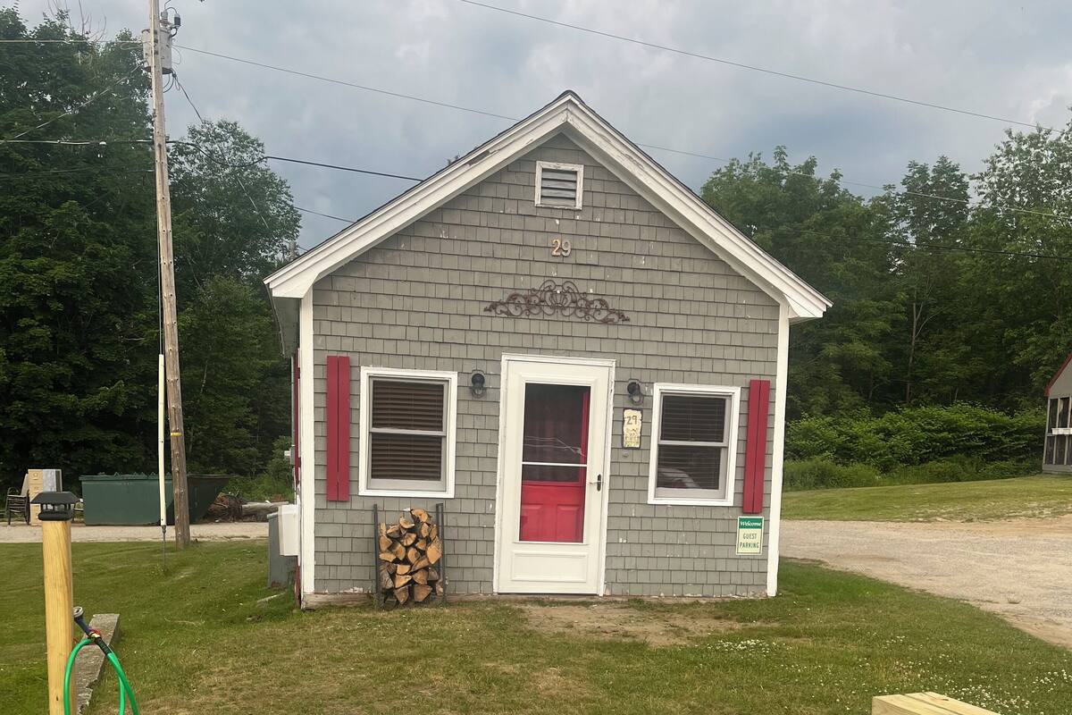 A small, single-story building with a red door and two windows with shutters, a fire pit in the foreground, and a bench to the right.