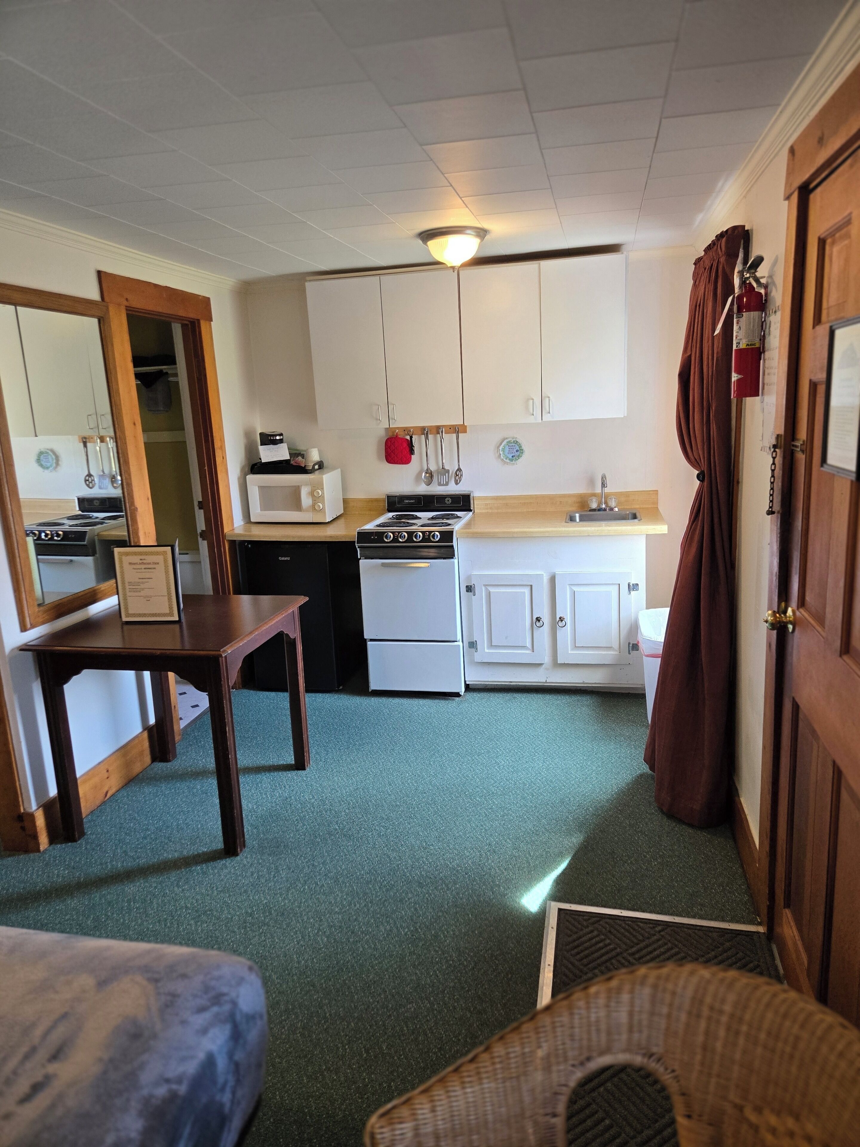 A compact kitchen with white cabinets, a stove, a microwave, and a sink. There is a wooden table and a wicker chair in the foreground.