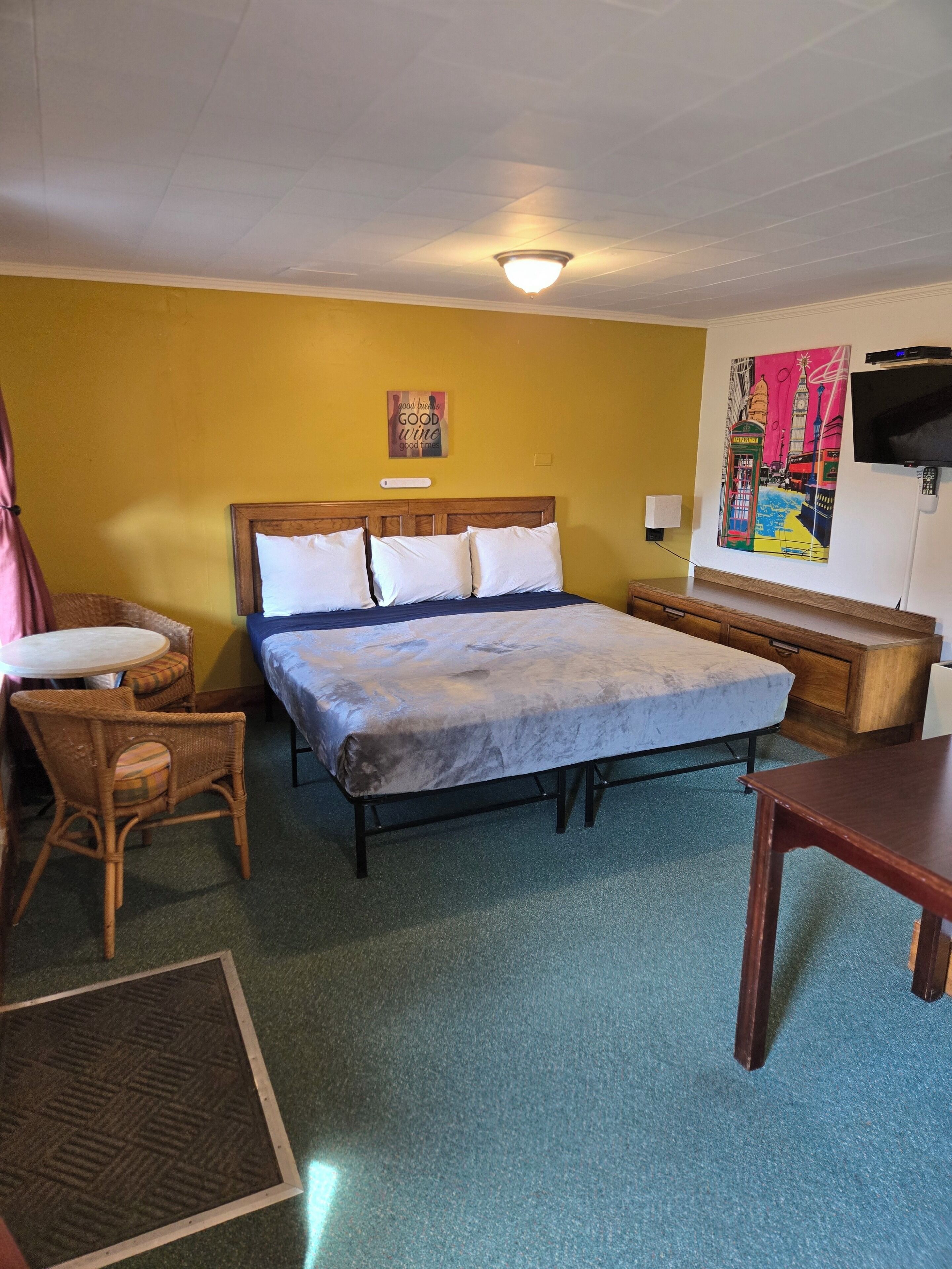 A hotel room with a bed, a chair, a small table, a TV, and a painting on the wall.