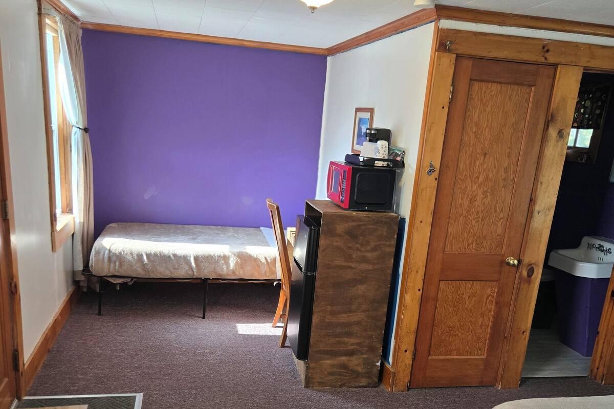 A compact room with a purple wall, a wooden door, a bed, a desk, and a small kitchenette area.