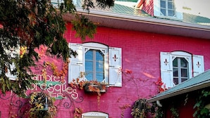 Un bâtiment en briques rouges, doté de volets blancs, d’une fontaine en pierre et d’un jardin où poussent différentes espèces de plantes.